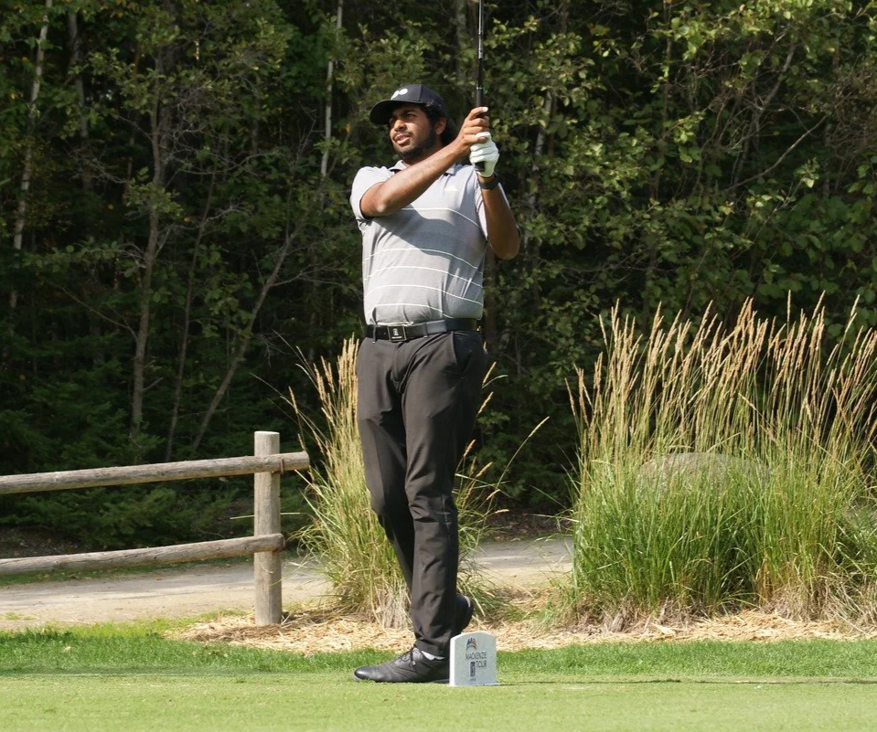 A man in a striped golf shirt and dark pants is on a golf course, holding a golf club after hitting a shot, with trees in the background and a wooden fence nearby.
