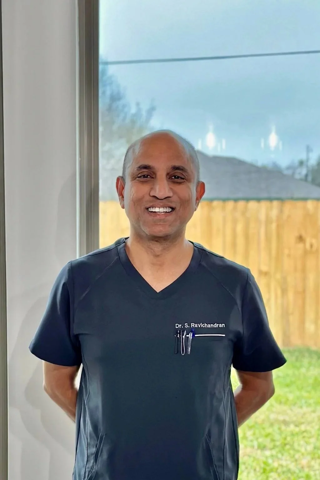 A smiling man wearing navy medical scrubs standing indoors near a window with a wooden fence and a house visible outside.