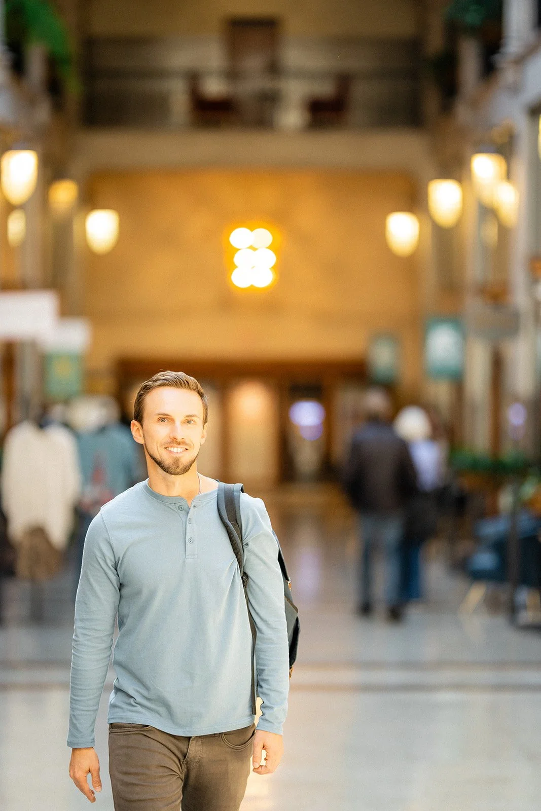 A smiling man with a backpack walking in an indoor public space, blurred background with people and warm lighting.