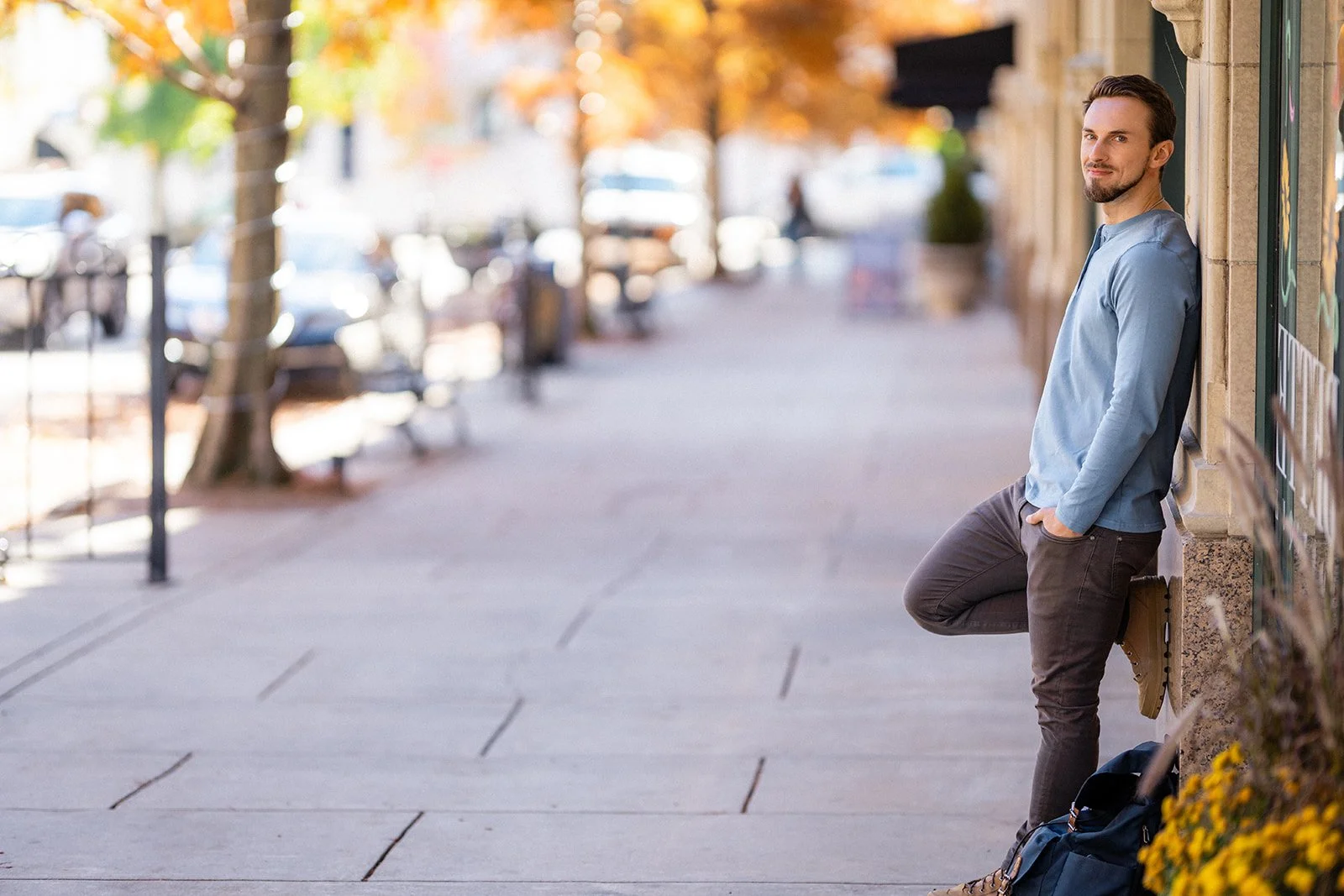 A man leaning against a wall on a sidewalk, looking at the camera with his hands in his pockets and a backpack on the ground beside him.