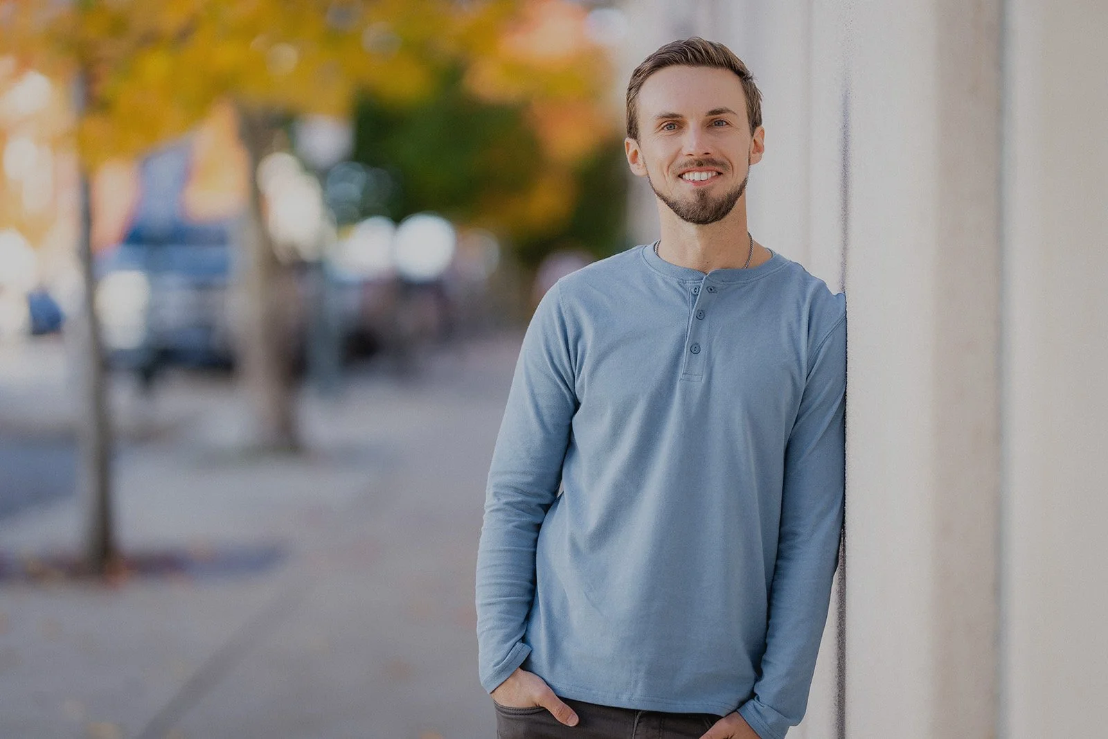A young man with light brown hair and a beard, wearing a light blue long-sleeve shirt, leaning against a wall outdoors with trees and cars blurred in the background.