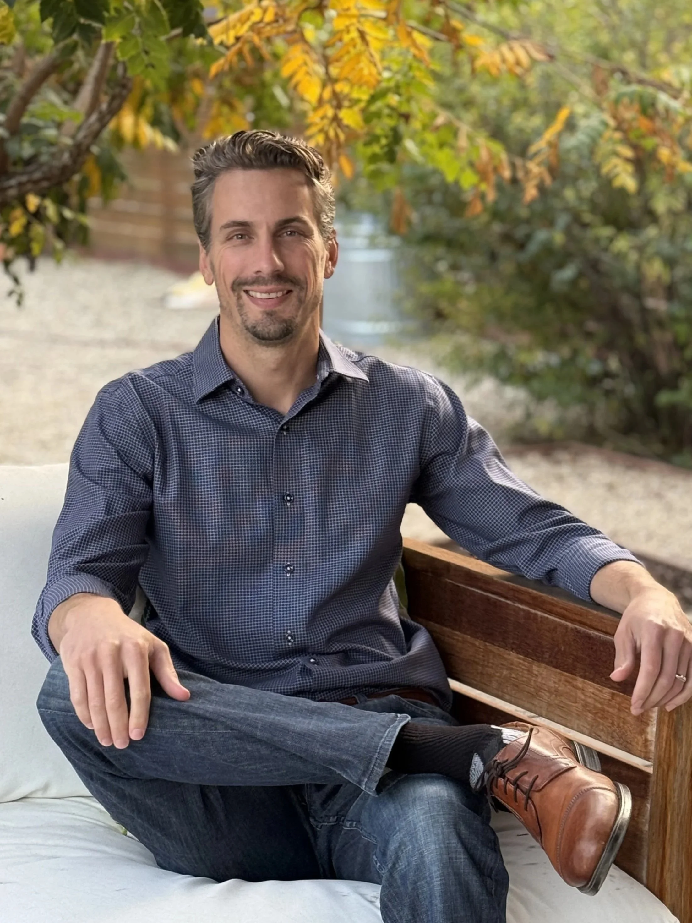 A man sitting on an outdoor couch with his leg crossed, smiling, wearing a checkered dark blue shirt, jeans, and brown shoes, with yellow and green leaves in the background.
