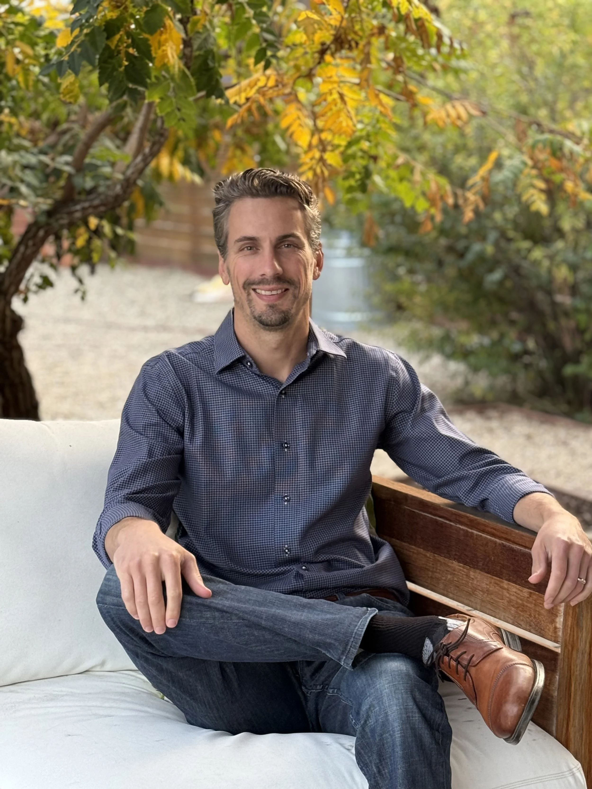 A man with short brown hair, a trimmed beard, and a mustache, sitting serenely on a white outdoor couch with wooden armrests. He's wearing a blue checked button-up shirt, dark jeans, and tan leather shoes, with one leg crossed over the other. The background features trees with yellow and green leaves, indicating an autumn setting.