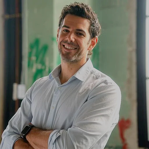 A smiling man with dark curly hair and a beard, wearing a white button-up shirt, standing indoors with arms crossed.