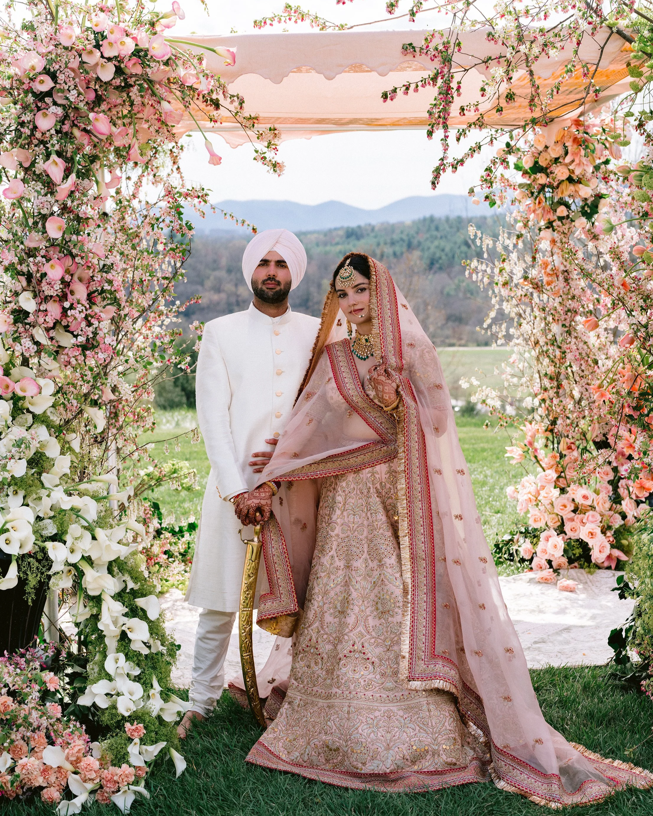 Indian bride and groom in traditional wedding attire, standing under a floral arch outdoors with mountains in the background.