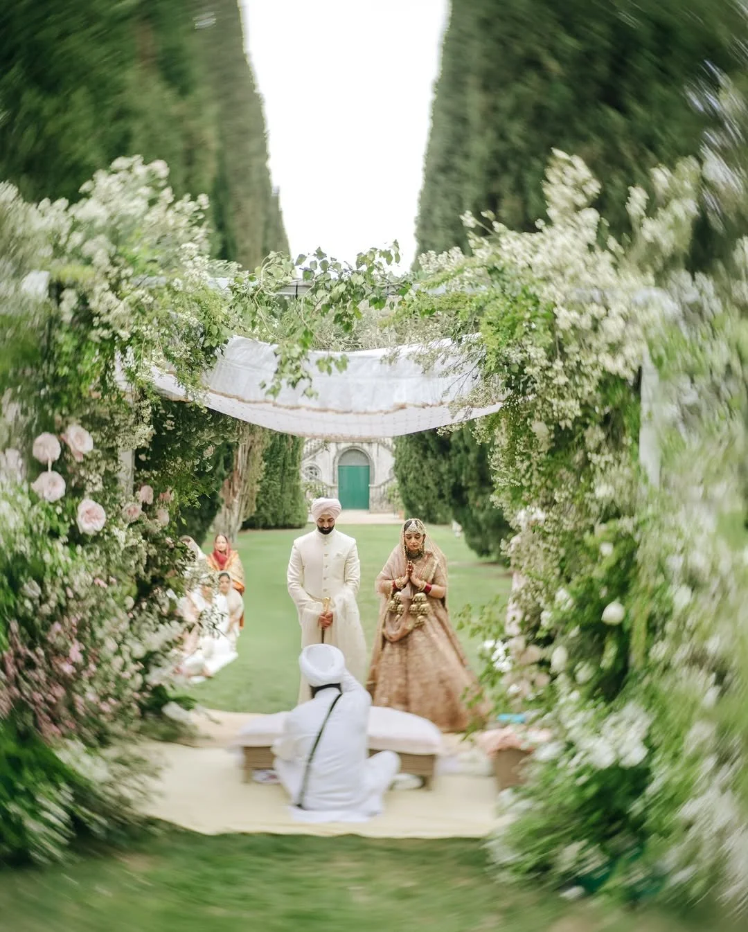 Indian couple performing a traditional wedding ceremony outdoors surrounded by floral decorations and greenery.