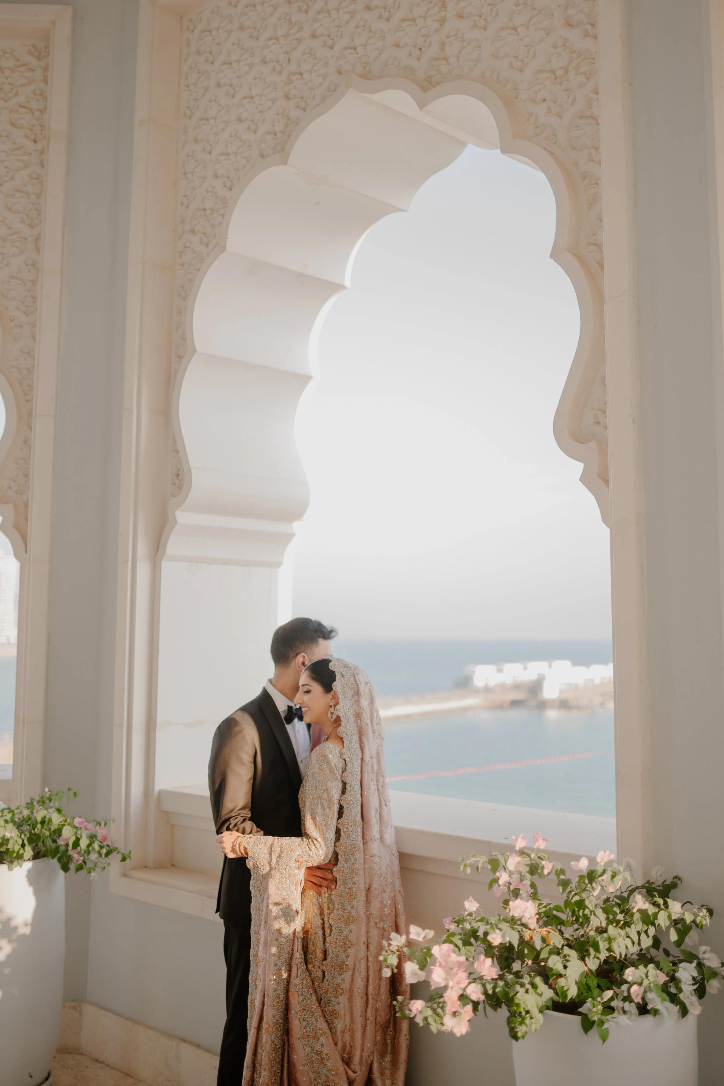 A newlywed couple in traditional wedding attire standing close together on a balcony, overlooking a body of water.