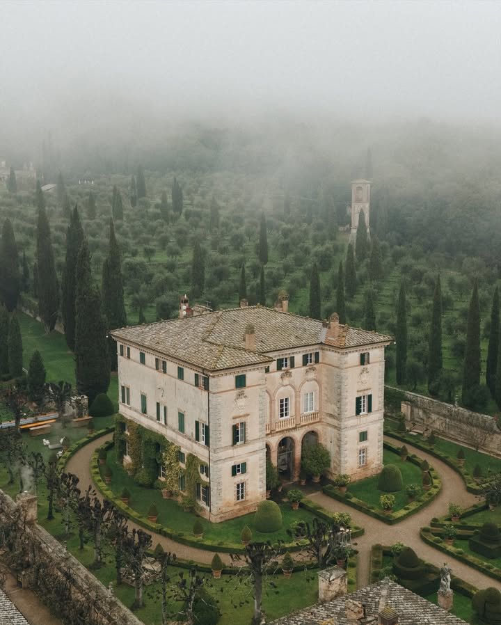 A large historic villa with a gravel driveway, surrounded by a well-manicured garden with trimmed bushes and trees, set against a backdrop of denseTuscan cypress trees and foggy hills.