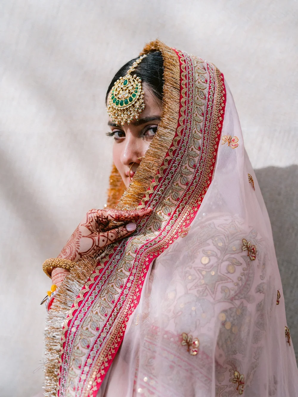 A woman dressed in traditional Indian attire with ornate jewelry, henna on her hand, and a pink embroidered saree with gold accents, partially covering her face.