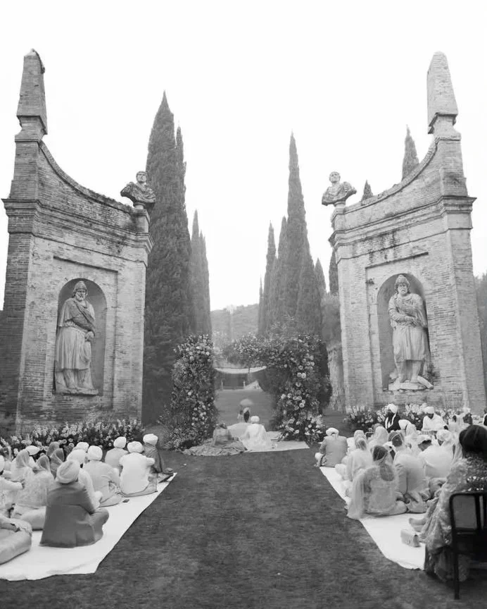 People sitting on mats in front of large brick entrance gates with statues and tall trees in the background, possibly attending an outdoor ceremony or gathering.
