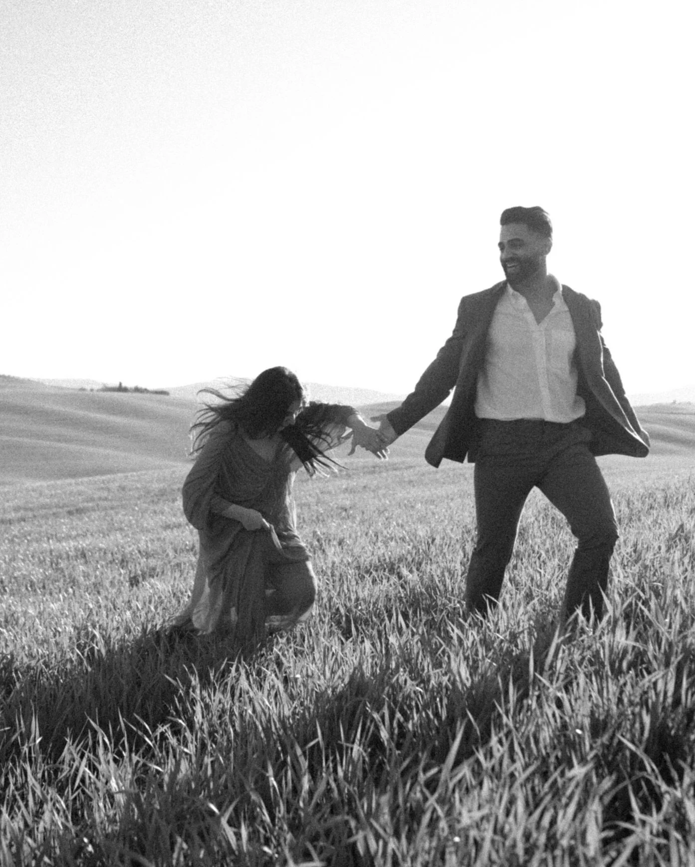 A black and white photo of a man and woman holding hands and smiling in a grassy field with rolling hills.