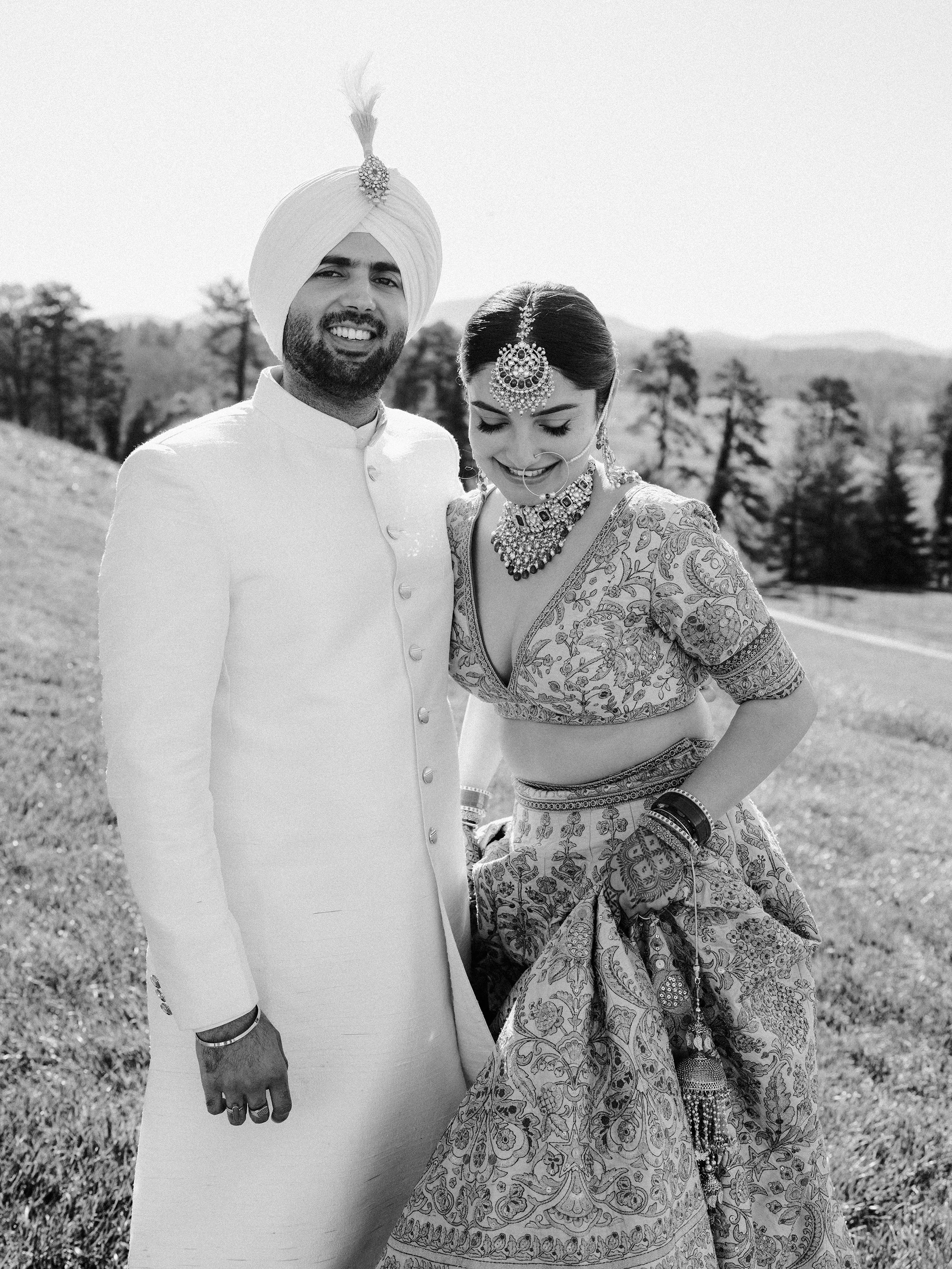 A couple in traditional Indian wedding attire standing outdoors on a grassy hill with trees and mountains in the background, smiling and looking down.