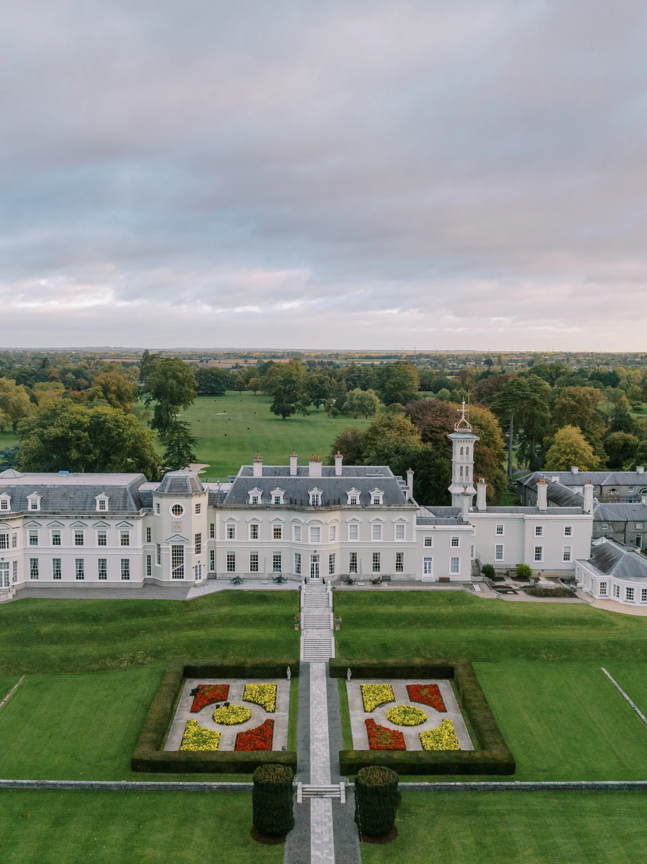 An aerial view of a large white mansion with a castle-like appearance, surrounded by neatly maintained green lawns and colorful flower gardens, set against a backdrop of a vast countryside with trees and open fields under a cloudy sky.