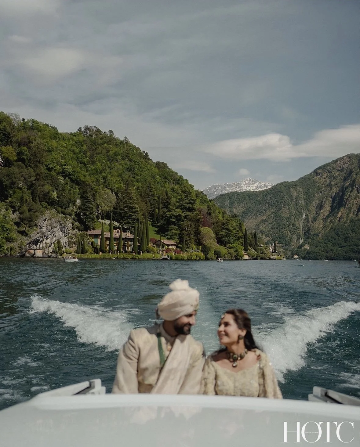 A couple dressed in traditional Indian attire on a boat, with scenic green hills and mountains in the background.