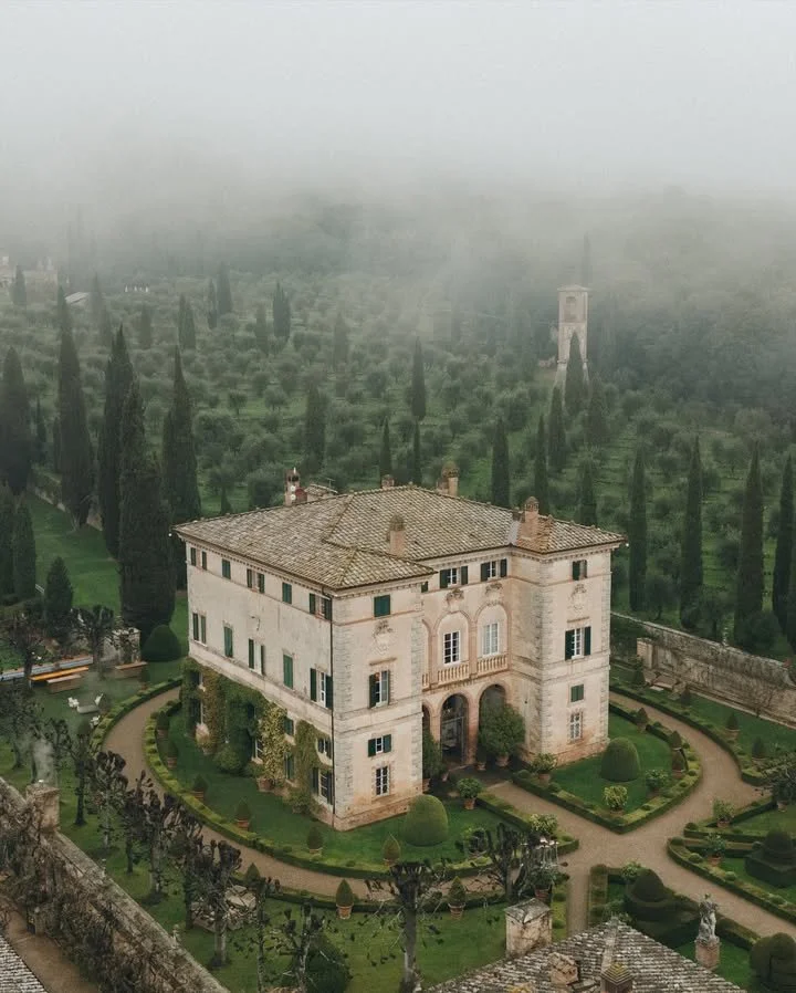 A historic stone mansion with a landscaped garden, surrounded by tall cypress trees, on a foggy day.