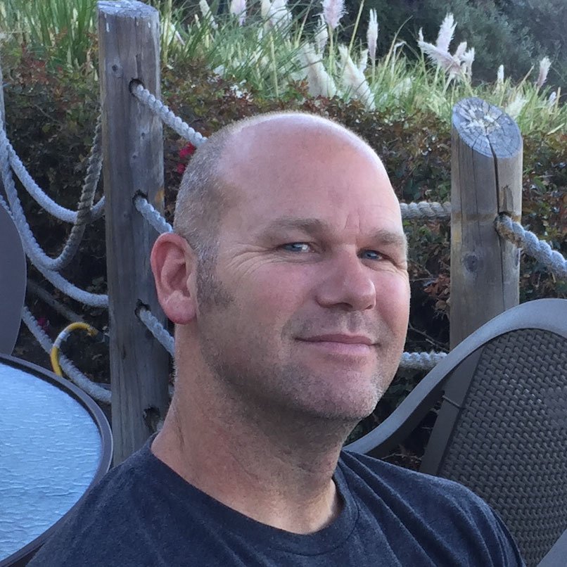 A man with a shaved head and light beard sitting outdoors near a wooden fence with rope railing. There is greenery and white ornamental grasses in the background.