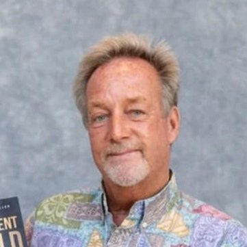 A man with light-colored hair and facial hair, wearing a colorful, patterned shirt, standing in front of a plain gray background.