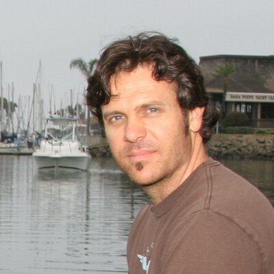 A man with dark curly hair and a slight beard standing near a marina, with boats and a building in the background.