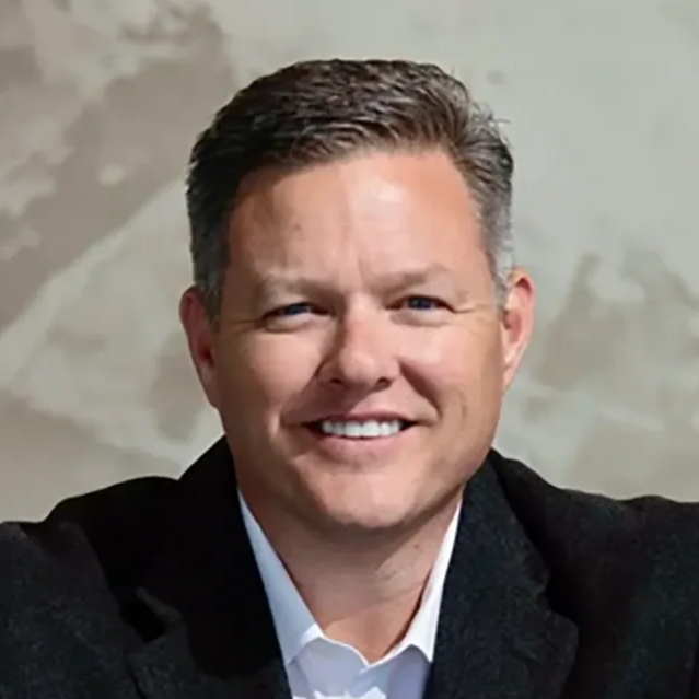 A portrait of a smiling man with short, dark hair, wearing a dark blazer and a white shirt, against a neutral background.