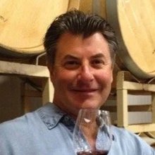 A man with dark hair smiling and holding a glass of wine in a winery setting with wine barrels in the background.