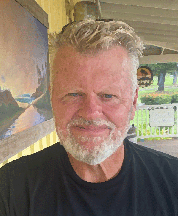 A close-up of a smiling older man with gray hair and a beard, wearing a black shirt, standing outdoors near a yellow building with a landscape painting on the wall.