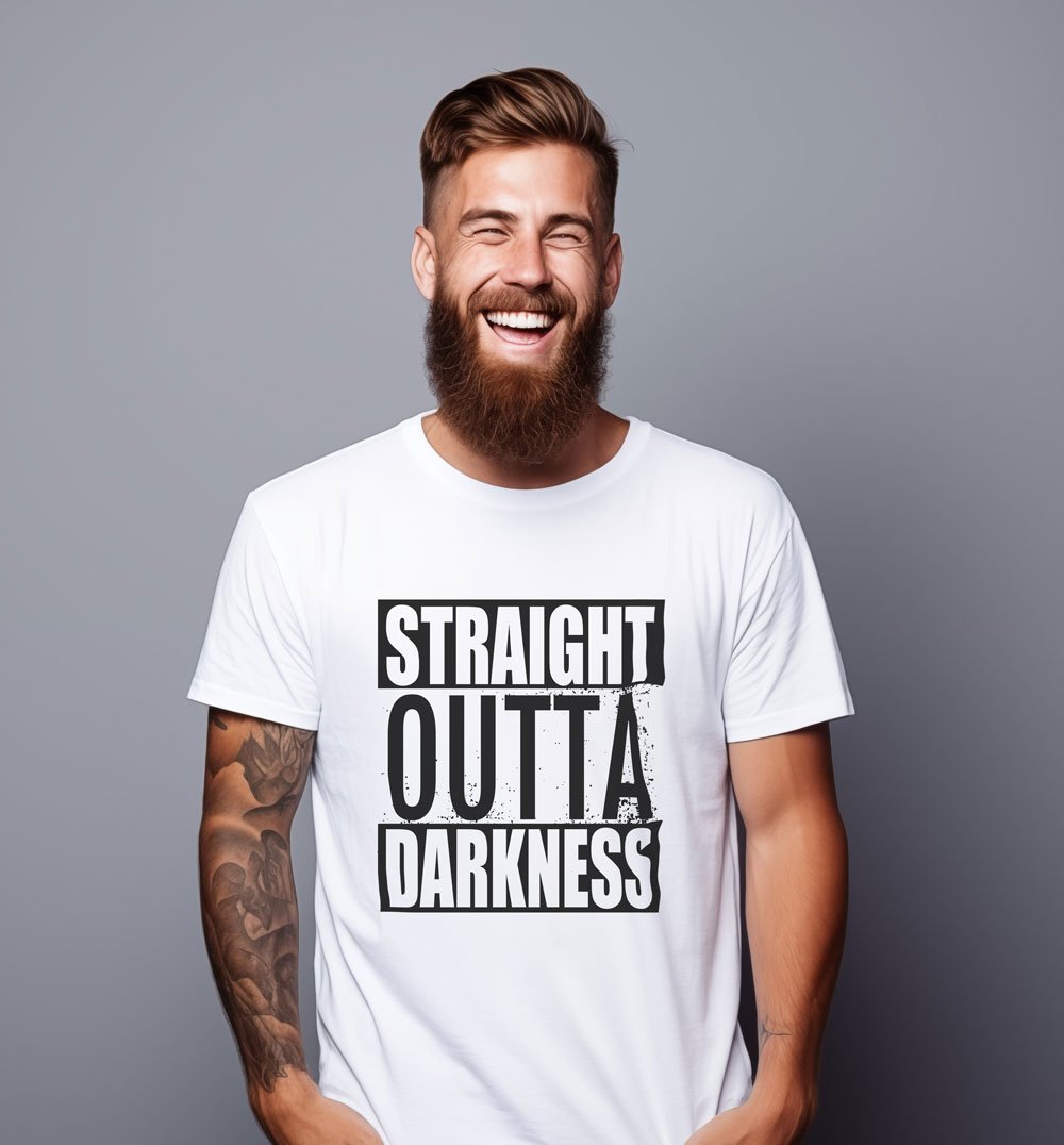 A man with a beard and tattooed arm smiling and wearing a white T-shirt with the text 'Straight Outta Darkness' on it, against a plain gray background.