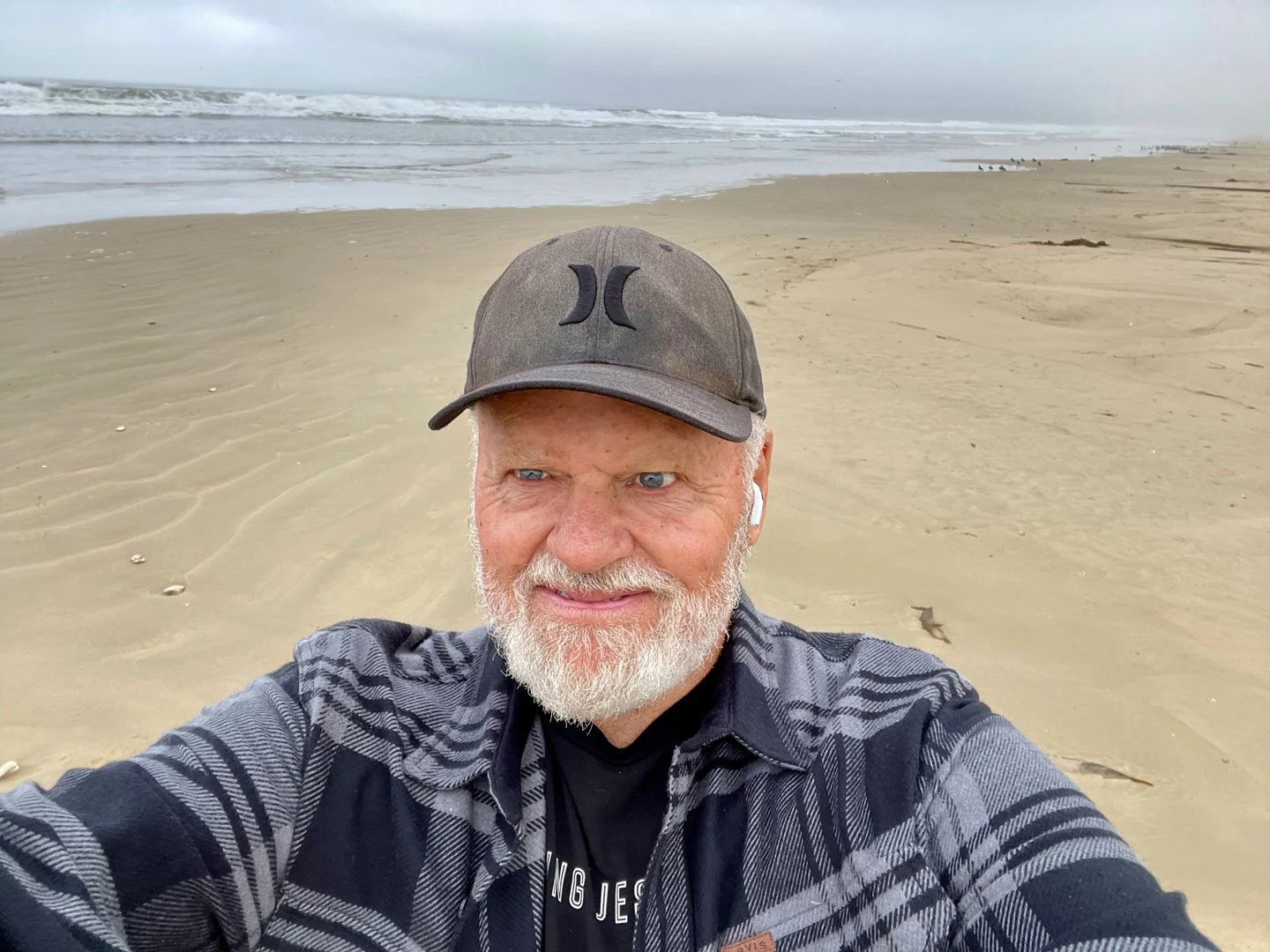 Scott Bailey wearing a gray cap on a beach, with the ocean and shoreline behind him.