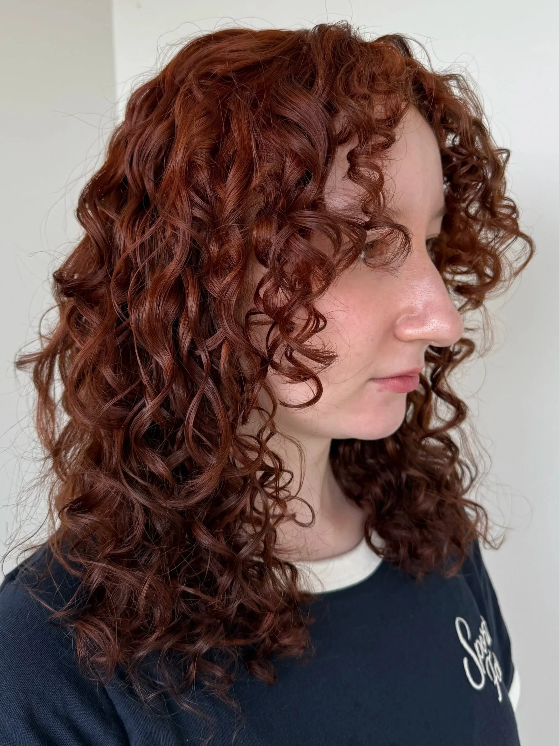 A woman with shoulder-length, tightly curled red hair, wearing a dark shirt with a white collar and the word 'Stay' embroidered on it, standing against a plain white wall.