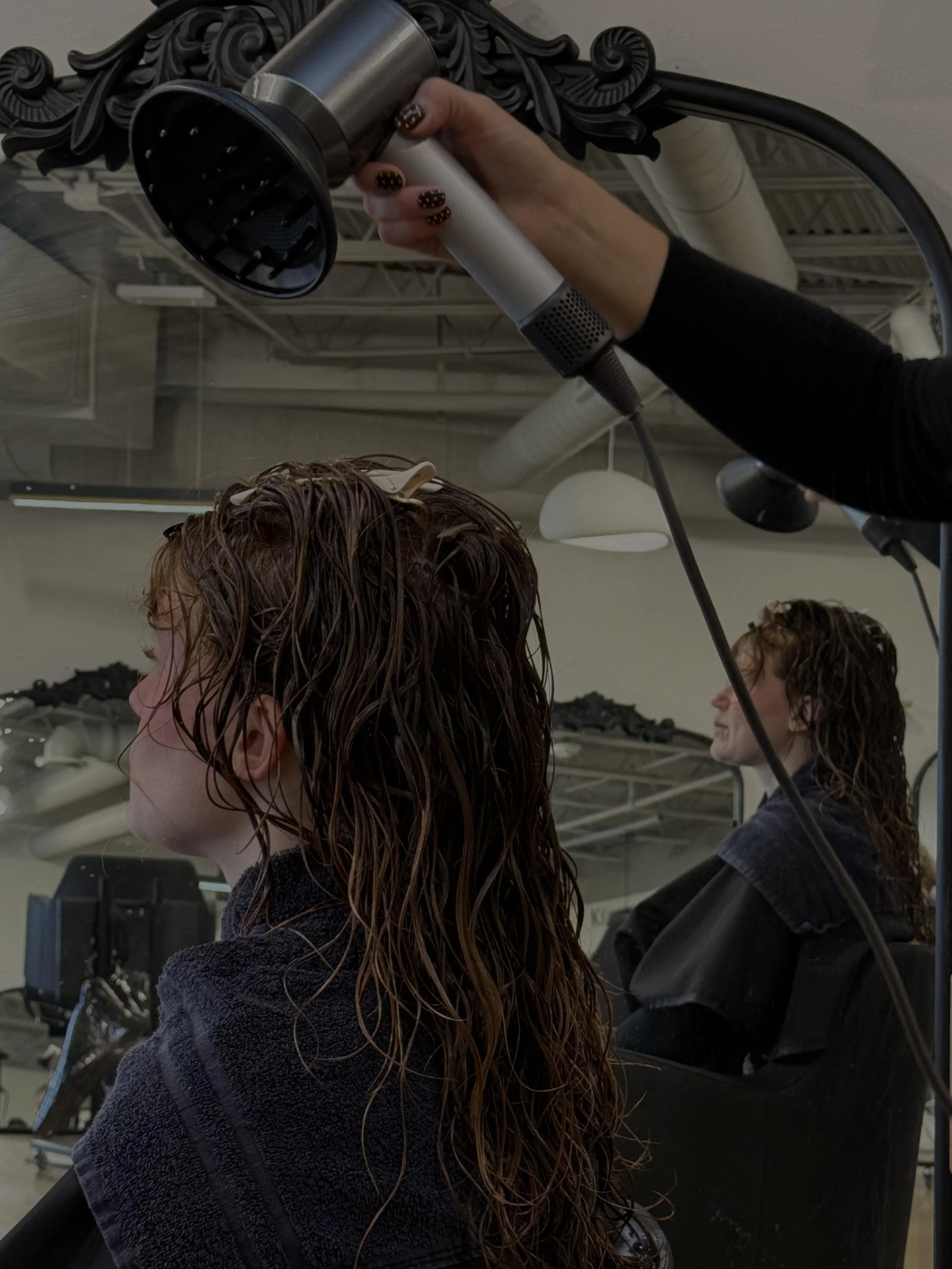 A person in a salon is blow-drying a woman’s wet, curly hair while she is seated in front of a mirror.