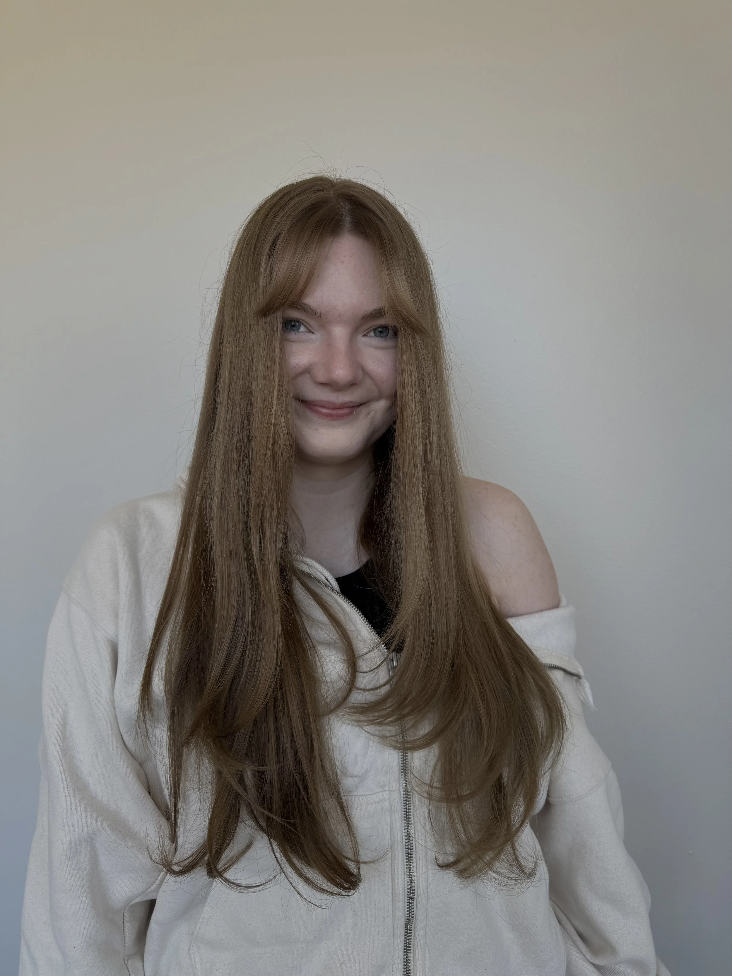 A young woman with long layered hair, wearing a white jacket off one shoulder, smiling at the camera against a plain wall.
