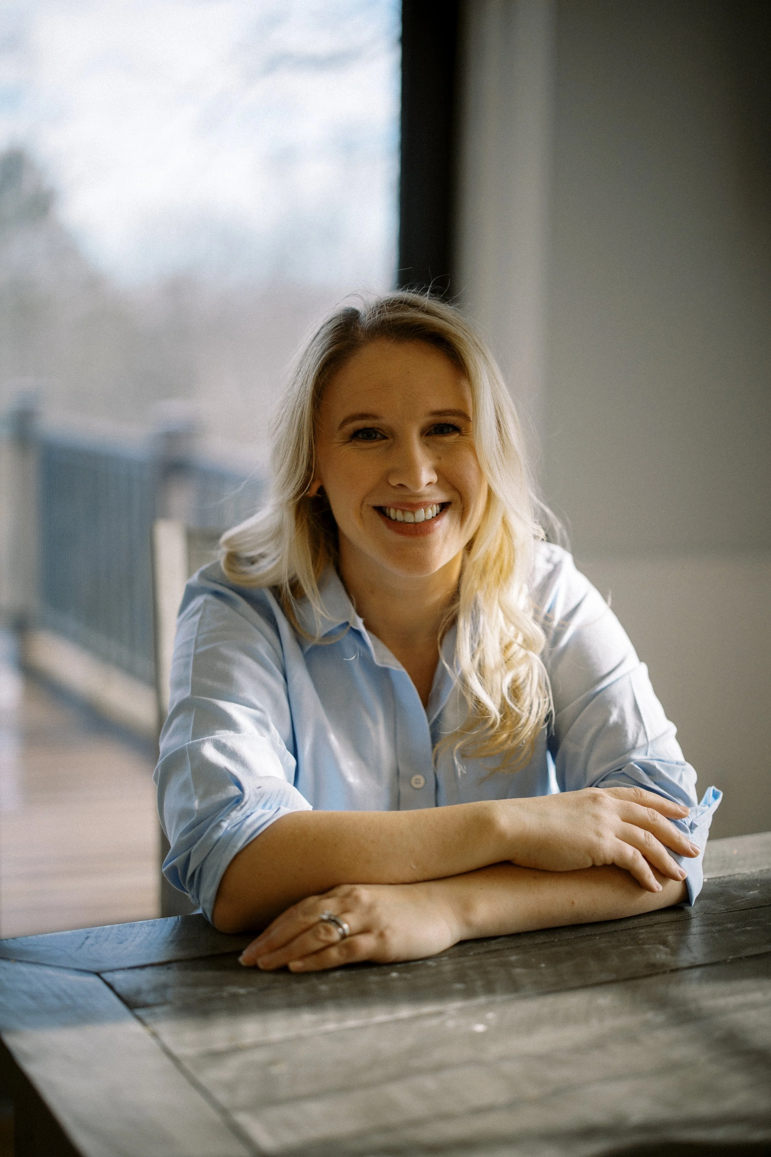 A smiling woman with blonde hair, wearing a light blue shirt, leaning on a wooden table inside a room with a large window showing an outdoor scene.