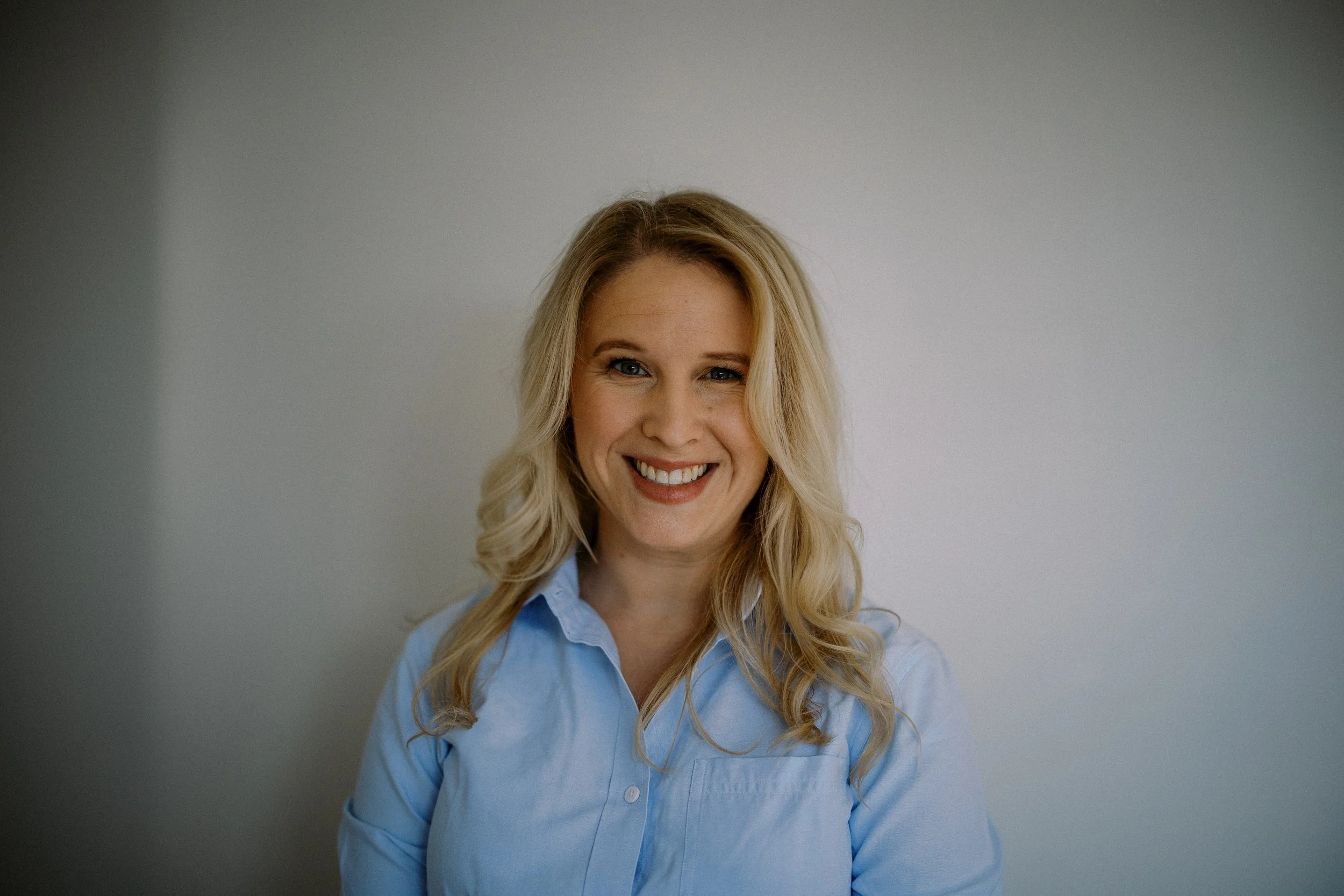 Portrait of a smiling woman with blonde hair wearing a light blue shirt, standing against a plain grey background.