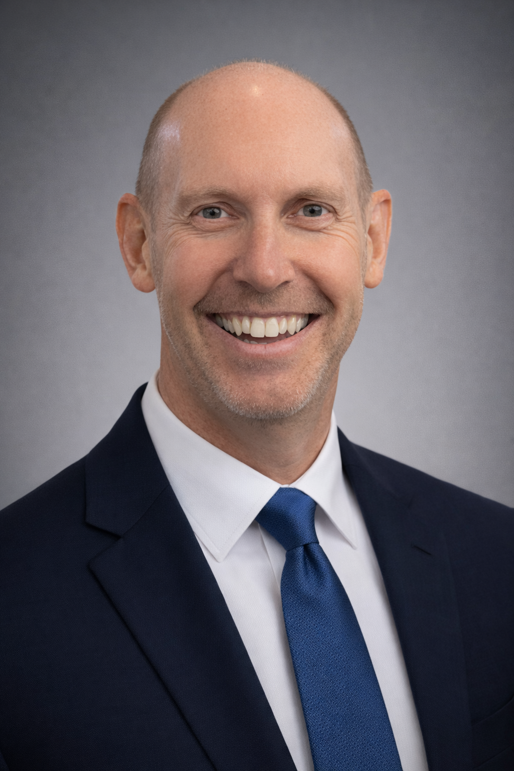 Professional headshot of a smiling man wearing a dark suit, white shirt, and blue tie, with a gray background.