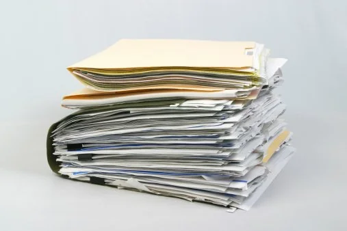 A large stack of disorganized papers and folders on a white surface.