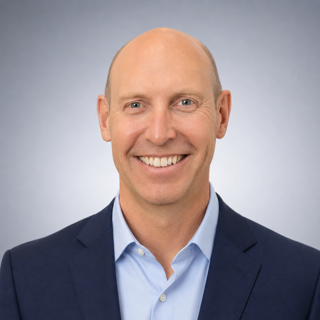 Professional headshot of a smiling man with a bald head, wearing a dark suit and light blue shirt, against a neutral gray background.