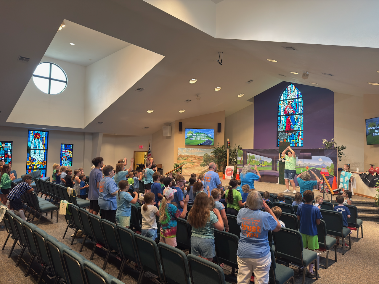 Children and adults in a church or community hall watching a stage performance with colorful stained glass windows, some with raised hands and following along with the presenter.