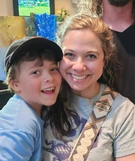 A smiling woman and young boy taking a selfie together inside an art studio with colorful paintings in the background.