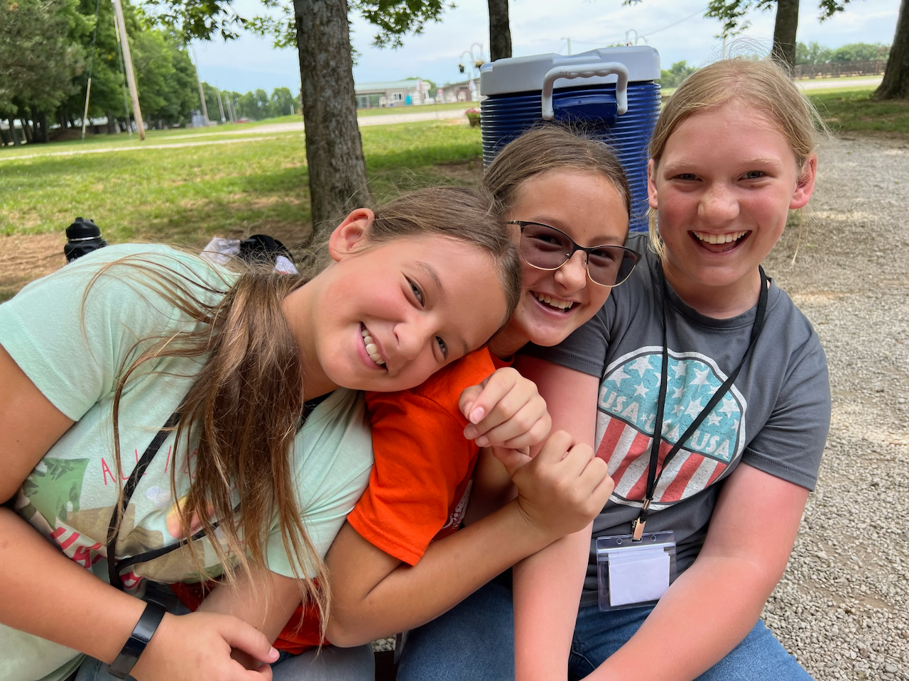 Three smiling girls embracing outdoors on a gravel area with trees and a grassy field in the background.