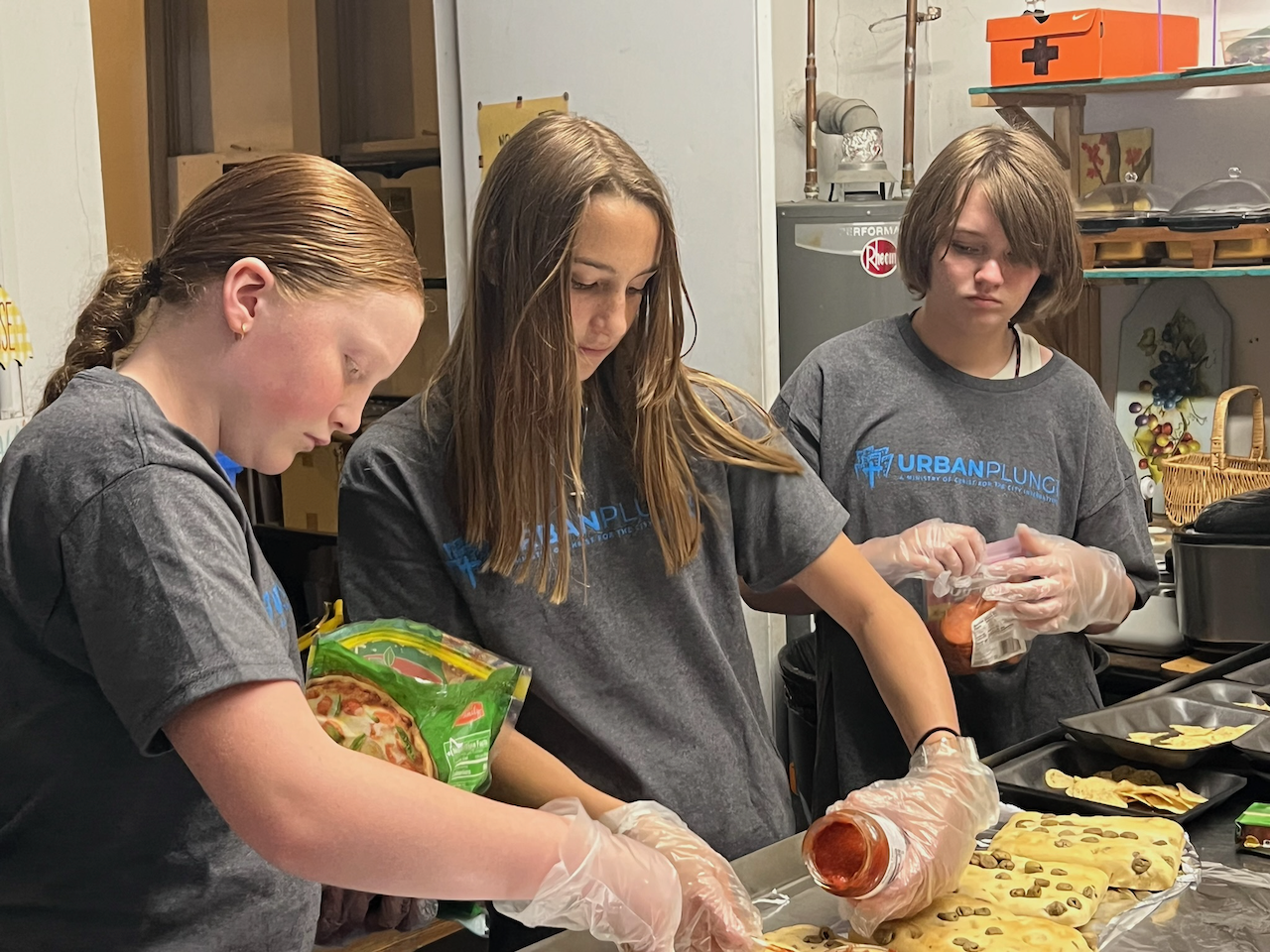Three young girls wearing gray T-shirts with 'URBAN PLUNG' written on them are preparing food in a kitchen. They are wearing disposable gloves and working on trays of cookies or pastries with chocolate chips. The kitchen has shelves with various items, a water heater, and a basket, with a focus on their activity of baking or food preparation.
