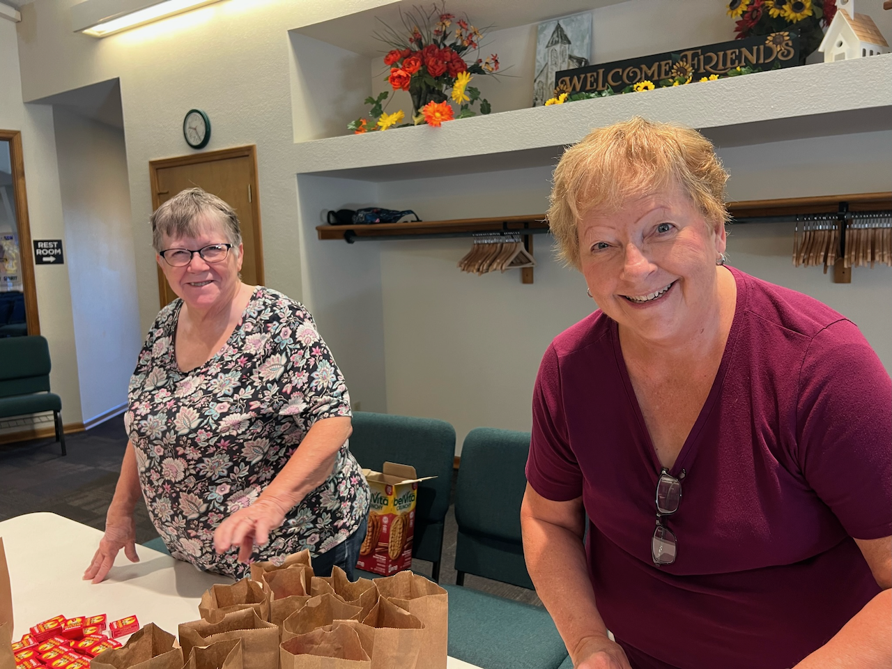 Two women smiling and packing small paper bags with snacks or small items at a table in a room decorated with flowers and a welcoming sign.