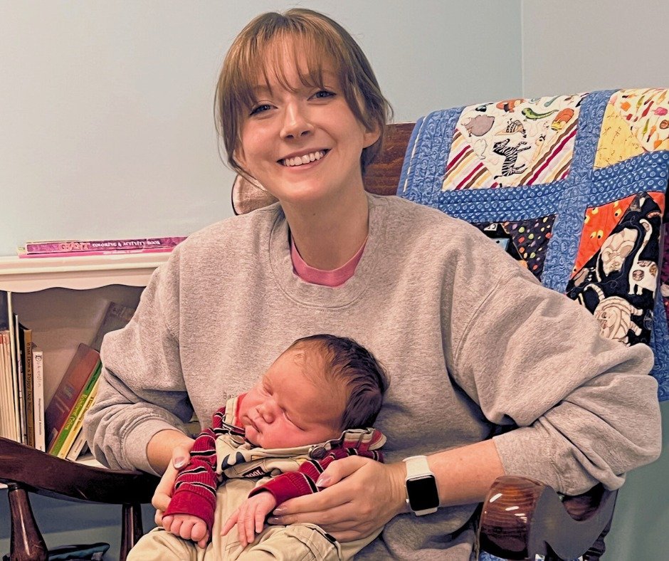 A woman sitting in a wooden chair, smiling and holding a newborn baby, with a colorful quilt hanging on the wall behind her.
