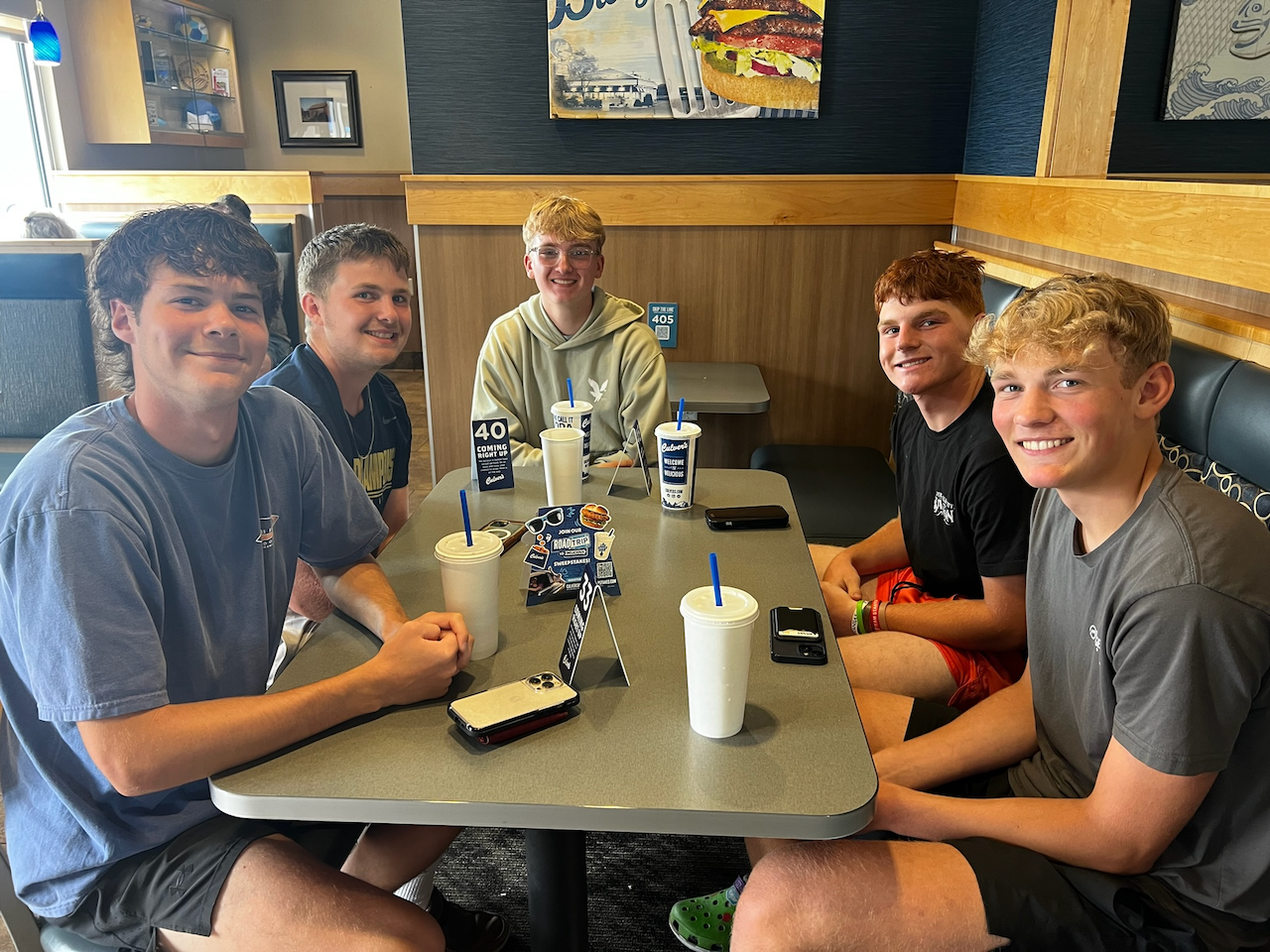 Six young men sitting at a table in a fast food restaurant, smiling at the camera, with drinks and phones in front of them.
