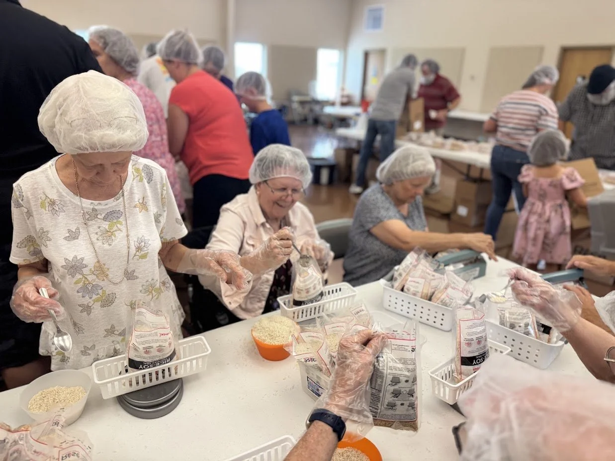 Older women packaging food in a community kitchen or food packing event, wearing hairnets and gloves, with several people working in the background.