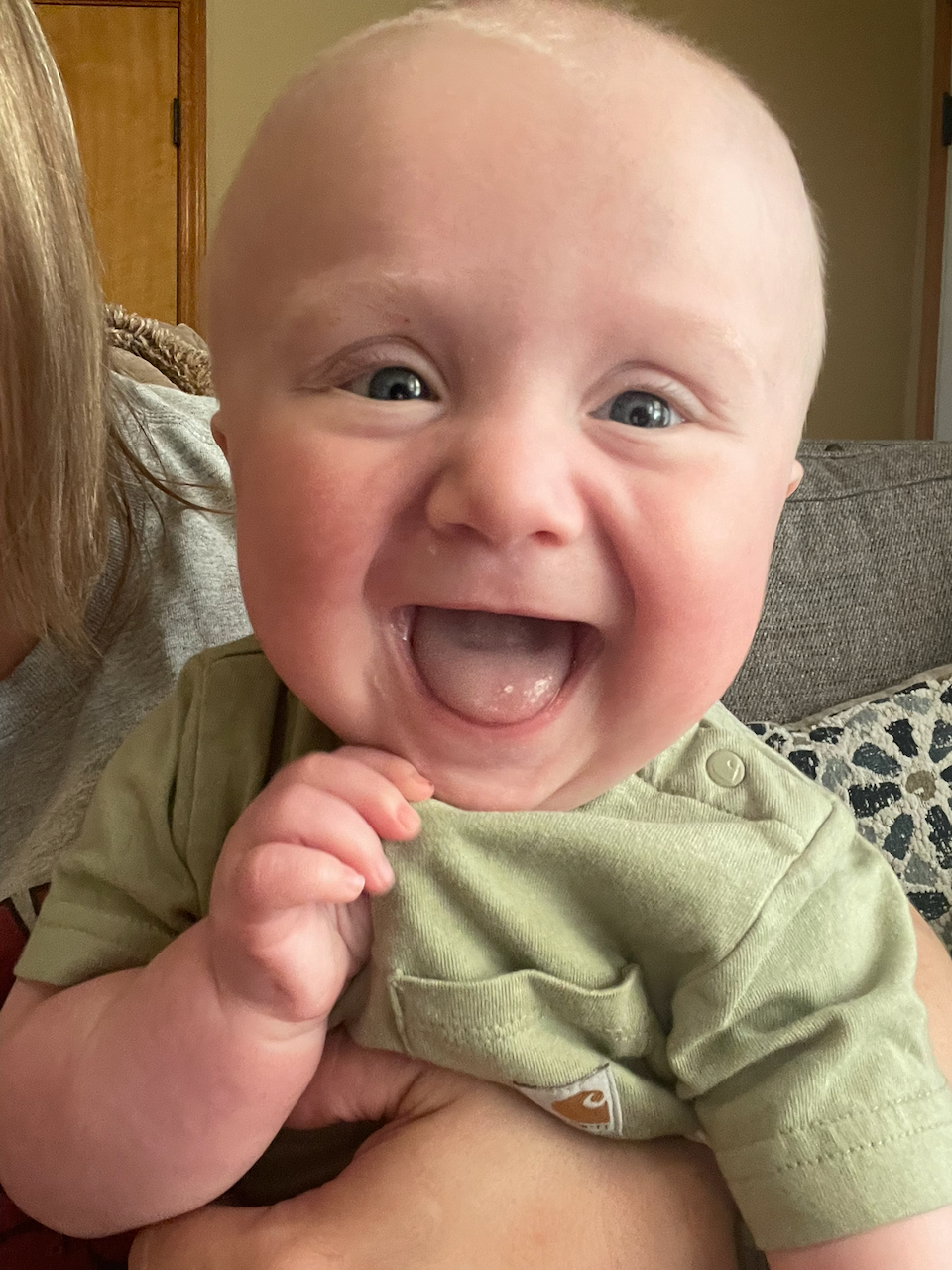 A happy baby with bright blue eyes, wide open mouth in a smile, and a bald head, being held by an adult.