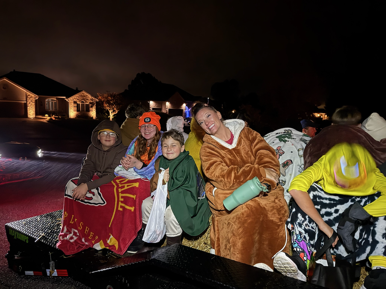 A group of people, including children and adults, riding on a trailer during a nighttime parade. They are dressed in casual and festive clothing, and some are wrapped in blankets or carrying water bottles. The background shows a suburban neighborhood under dark skies.