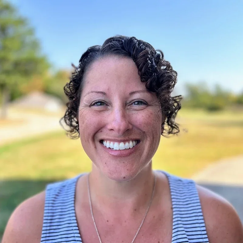 A woman with short, curly dark hair smiling outdoors on a sunny day, wearing a blue and white striped sleeveless top.