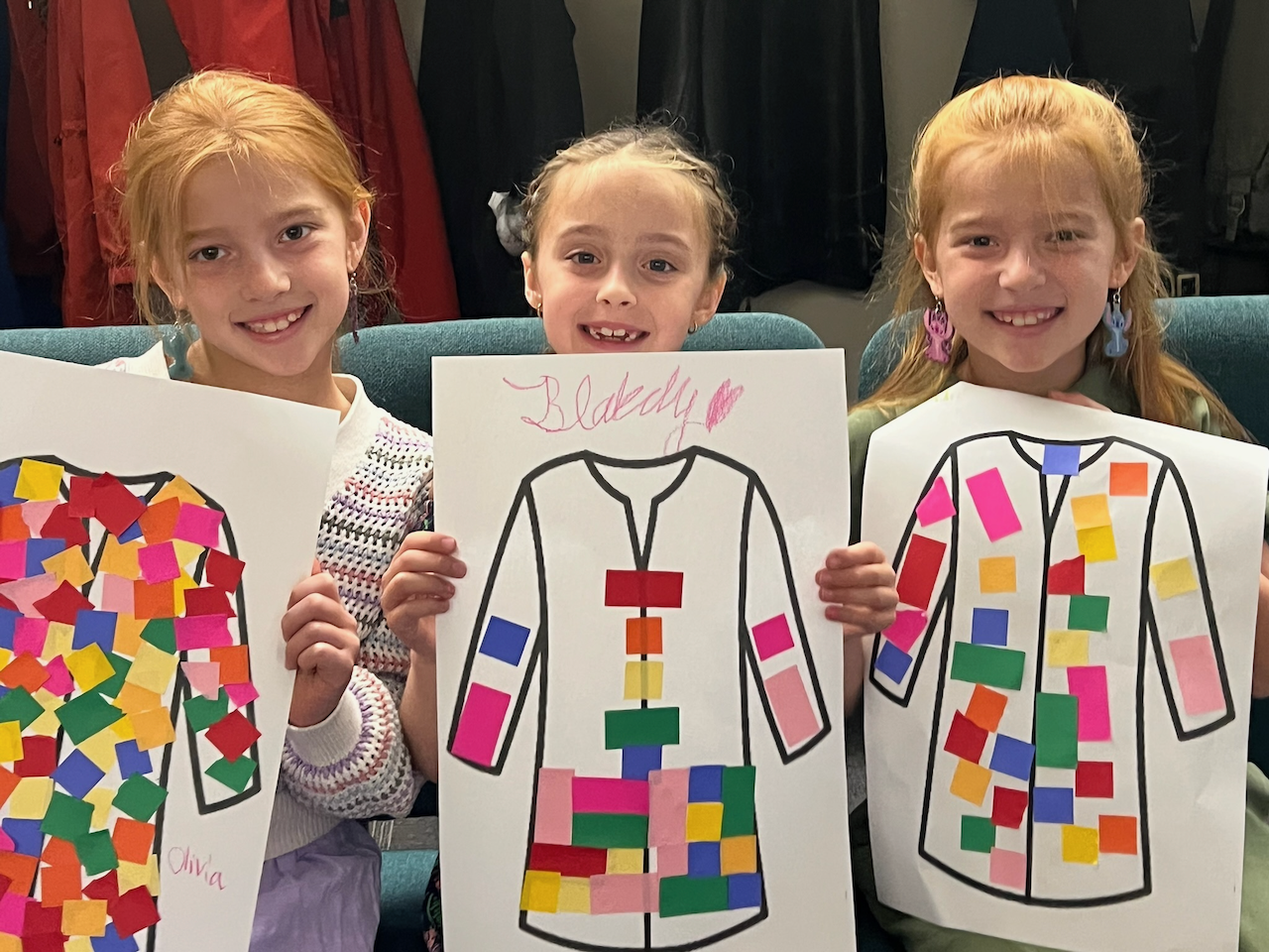 Three young girls standing together, holding large drawings of coats outlined with black lines, filled with colorful sticky notes, in a room with hanging coats in the background.
