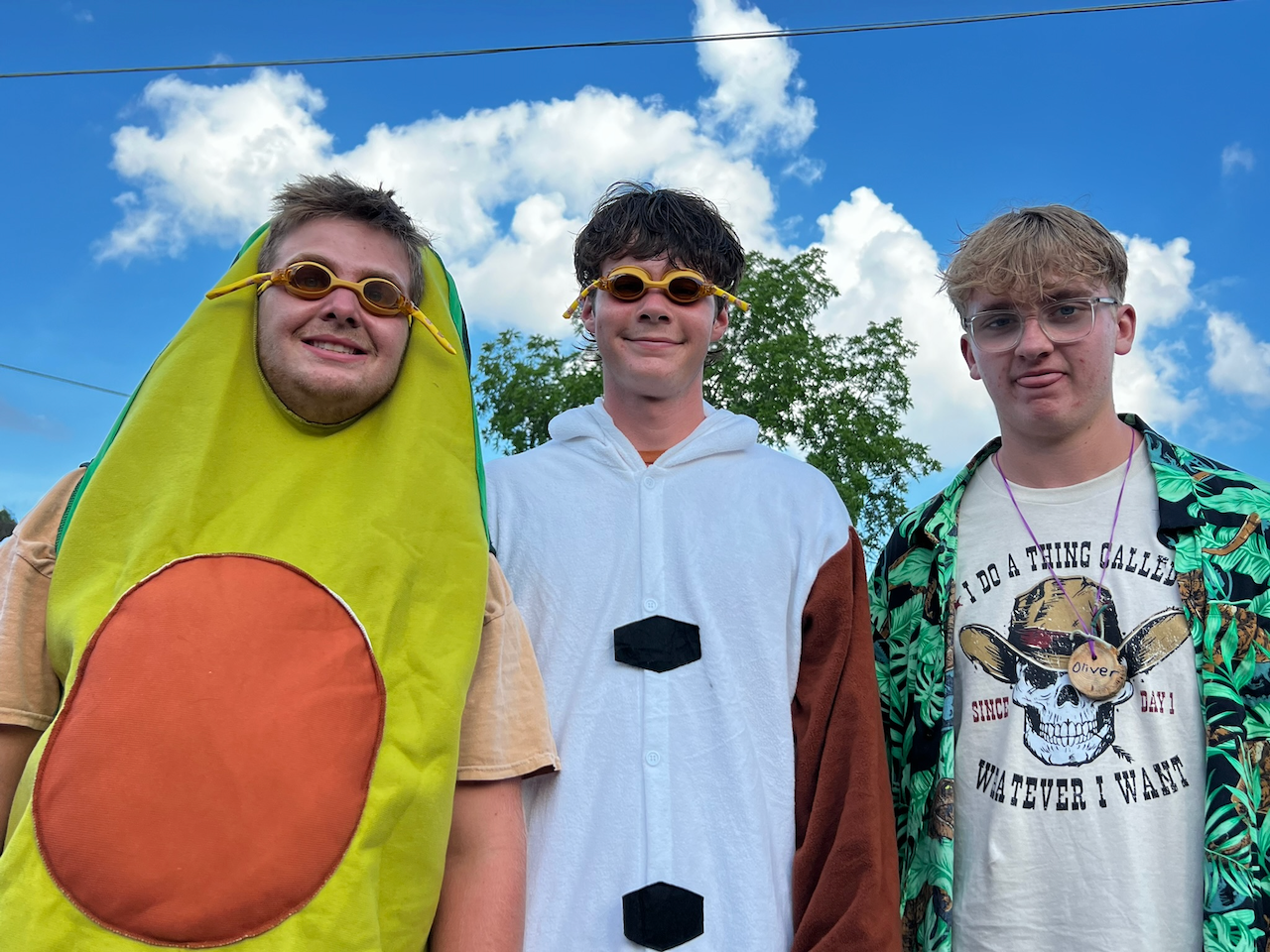 Three young men standing outdoors under a blue sky with clouds, wearing playful costumes and sunglasses, smiling at the camera.