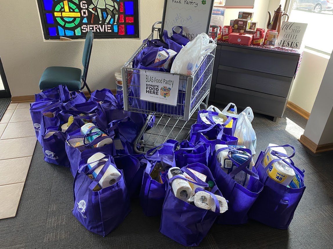 Multiple purple tote bags filled with food items and supplies, placed around a shopping cart labeled 'BLAS Food Pantry' inside a room with a stained glass window and a cabinet with shelves on top.