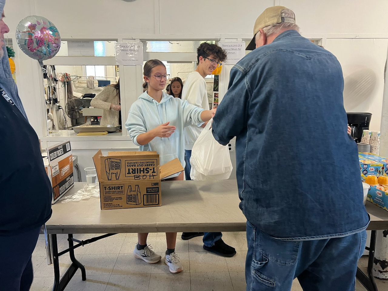 People at a food distribution table, two young women and an older man, with boxes and supplies in the background.